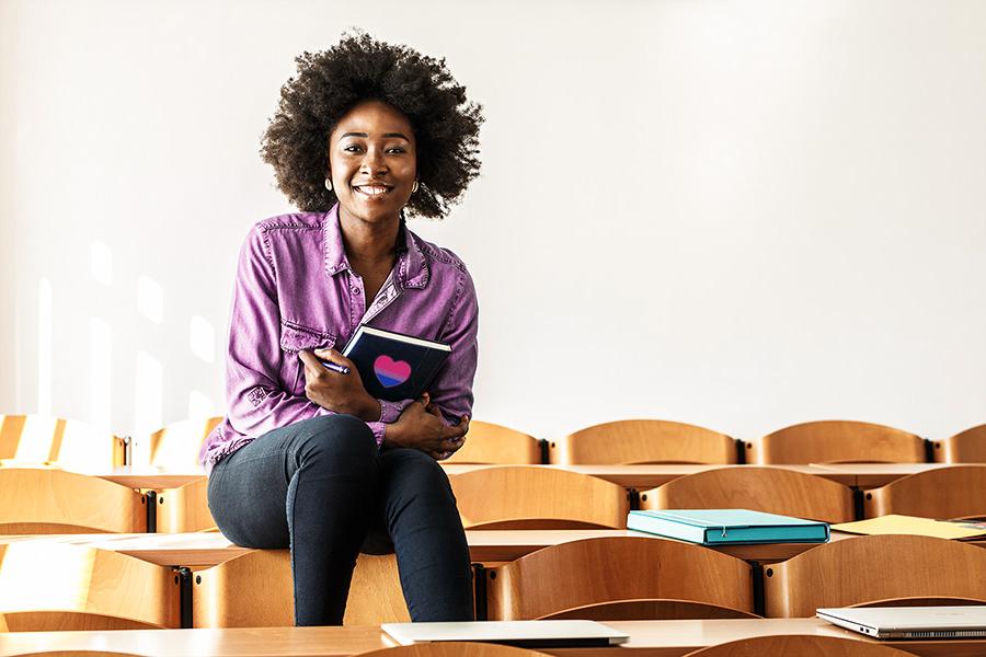African American student sitting in college classroom holding a notebook that has the bisexual flag shaped like a heart on it
